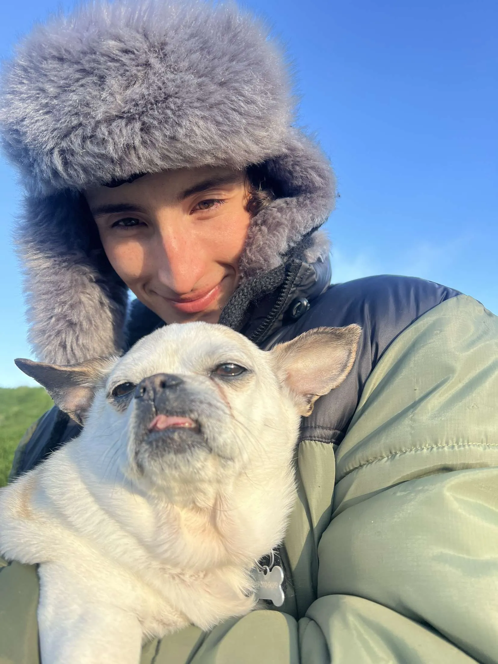Jackson in a grey fur lined trapper hat and a puffer jacket outdoors under a clear sky, holding a small white chihuahua against his chest
